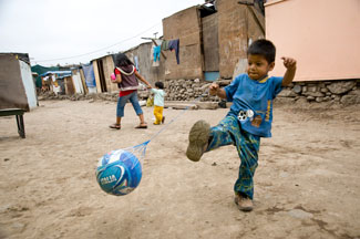 Young boy hangs on a chain link fence in front of tents in a refugee camp.