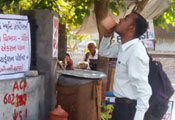 In India, a man drinks from a cup at a public outdoor drinking water station in the shade on a street corner