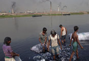 Many men wading in river bank rinsing large clumps of plastic bags, smoking factories in background on opposite bank