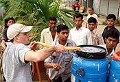 In Dhaka, Dr. Eric Nelson pumps water through a hose from a pond into a blue barrel, surrounded by others who are helping.