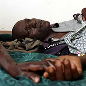 Man lying down in visible pain, his hand is held, in a hospital in Rakai, Uganda.