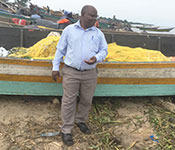 Dr. Joseph K.B. Matovu stands in front of a fishing boat on the shoreline in Uganda, many boats in the background.