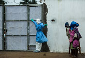 Photo by Morgana Wingard, courtesy of USAID, Healthcare worker outside Ebola clinic in full protective gear holds gate closed, warns away approaching people