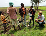 Two global health students collect data on mobile devices from community members, rural farmland in the background.