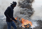 Man prods pile of burning electronic waste with a stick. Fire gives off a large cloud of thick, dark smoke.