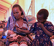 Photo by UNICEF Ethiopia/2021/Nahom Tesfaye. Woman holds a newborn baby, older child seated next to her on a cot, in a large tent clinic.