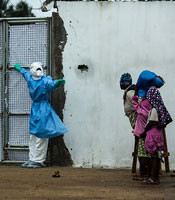 Healthcare worker outside Ebola clinic in full protective gear covering holds gate closed, warns away approaching people