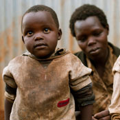 Young child in very dirty clothes, parent seated in background looking down