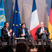 Roger Glass seated on a stage during the World Health Summit with two other men, multiple flags in the background