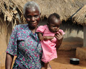 Older woman holds baby outdoors, thatched huts in the background