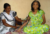Older woman seated cross-legged on bed, looks at camera while medical worker takes her blood pressure