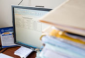 Stacks of paper on desk next to computer monitor showing data