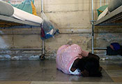 Photo by Torsten Blackwood / AFP / Getty Images. Person lies on floor curled into ball facing away from camera next to metal bed frames in an institution.