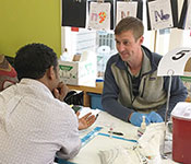 Photo courtesy of Dr. Rena Patel/project Harambee. Healthcare worker sits across from health fair participant, testing supplies on the table between them.