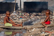 Two young, shirtless boys seated outdoors surrounded by garbage and rubbish, and stagnant water, background air is hazy