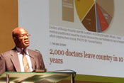 Dr. Nelson Sewankambo speaks into microphone at podium, slide reads 2,000 doctors leave country in 10 years