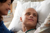 Young man in hospital bed wearing gown, with tubes in nose and no hair, female medical workers stands on each side