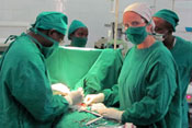 Dr Robin Petroze in green hospital scrubs and mask during a surgery with one additional surgeon, two workers in background