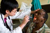 Woman doctor in white coat wears stethoscope, holds male patient's head back, uses pen light to look in his mouth