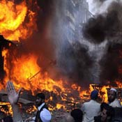 Bright orange flames surrounded by dark black smoke in a city street, buildings in background, people in foreground