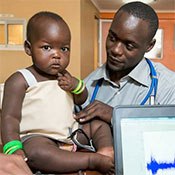 Photo courtesy of Mama-Ope. Medical worker examines young child who is sitting on an examining table in an indoor clinic setting, computer in foreground.