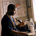 Researcher wearing scrubs in lab prepares samples, microscope in background, poster on wall reads Malaria