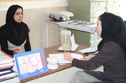 Two women, both in black headscarves, sit across from each other. One, facing the camera, speaks while gesturing with her hands