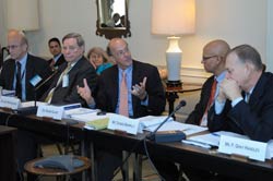 PHOTO: Dr. Roger Glass gestures with his hands while speaking, seated at a long table behind a microphone, 4 other men seated