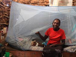 Bed net covers a bed, woman seated on edge of bed lifts the edge to show child lying down