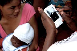 Woman holds a young baby in the background while a woman in the foreground enters data on a handheld device