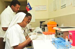 Two women and one man medical worker prepare vials and review notes at counter, Haitian flag hangs on wall