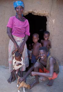 Photo: African woman stands in front of entrance to a hut, five children of varying ages surround her, one child pets a puppy