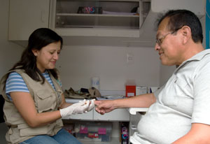 Photo: Dr Magaly Blas, seated in an exam room wearing gloves, collects a blood sample from the finger of a man seated opposite