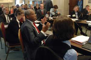 Photo: Meeting room full of attendees, many seated around a U-shaped table covered with papers, microphones and computers