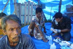 Photo: In a blue tarp tent a man in foreground stares vacantly, men seated in the background receives care from a man kneeling