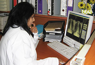 Photo: Dr Naidoo in a white lab coat seated at a desk reviews side by side chest x-rays on a computer screen