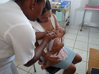 Photo: in exam room woman seated holds baby in her lap, nurse standing administers oral vaccine