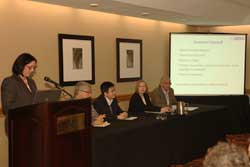 Photo: in a hotel conference room a woman stands behind a podium next to a long table with three men and one woman seated