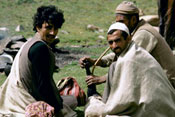 Three Indian men sit on the grass in a field, one smokes a water pipe, horses in background
