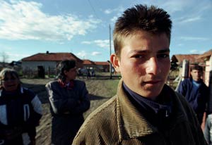 Young Bulgarian man looks into camera, village buildings and other people in background