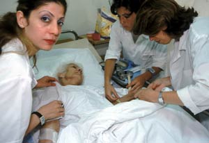 Older woman in hospital bed, three women in white jackets stand around her bed, adjusting her arms, one woman looks at camera