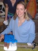 Dr. Julia Kubanek seated at wooden table smiles at camera, several specimen collection tubes stacked in holder on table
