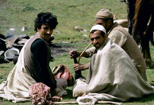 Three Indian men sit on the grass in a field, one smokes a water pipe, horses in background