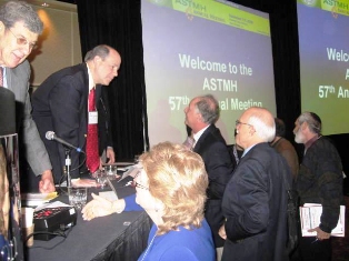 People greeting the lecturers of the symposium with two screens announcing the ASTMH meeting in the background
