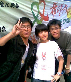 two Chinese male volunteers make a victory sign while a young girl poses with them. A sign with Chinese writing is in the background.