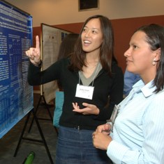 A woman points to a bulletin board while speaking to a woman