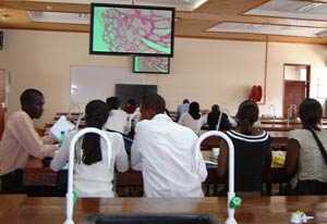 Students sit at desks in classroom, man sits behind table at front of classroom, addresses students