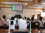 Group of students seated in modern, light-filled lab classroom view images on white screen monitors suspended from ceiling