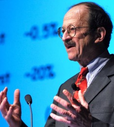 Dr. Harold Varmus speaking with a turquoise-colored slide behind him showing two percentages