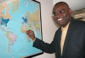 Dr. Clement Adebamowo looks at camera, smiling, points to Nigeria on a world map